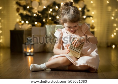 Excited curious little girl smiling, opening christmas gifts. Beautifully decorated christmas tree and house with lights and lantern. 
