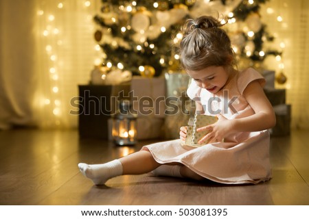 Excited curious little girl smiling, opening christmas gifts. Beautifully decorated christmas tree and house with lights and lantern. 