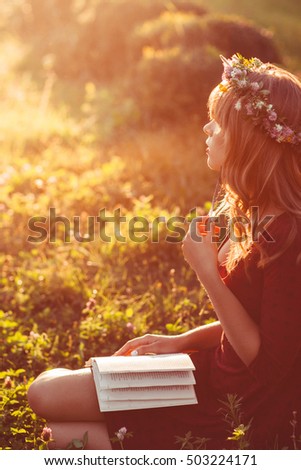 Profile of girl in wreath with book, sun flare effect. Free space on green meadow, attractive woman relaxing in nature during sunset. Rest, education, unity with nature concept