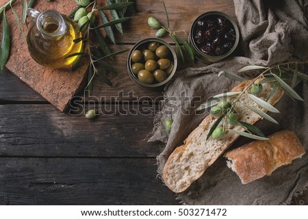 Green and black olives in tin cans with loaf of fresh bread and young olives branch on sackcloth, bottle of olive oil on clay board over old wood background. Overhead view with space for text Royalty-Free Stock Photo #503271472