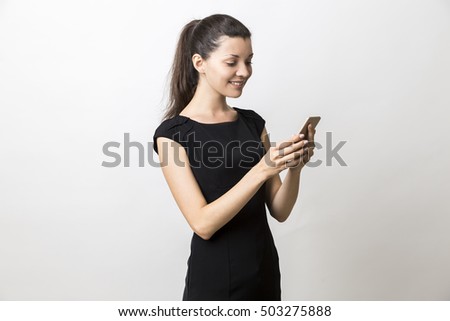 Portrait of young lady in beautiful black dress looking at her cell phone screen and standing against white background. Mock up