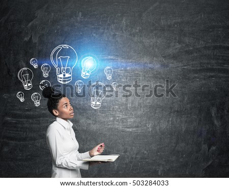 Side view of African American girl with notebook standing near blackboard with light bulb sketches. Concept of new idea. Mockup