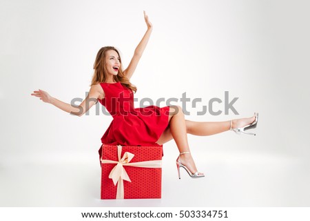 Cheerful attractive woman in red dress having fun while sitting on the big gift box isolated on a white background