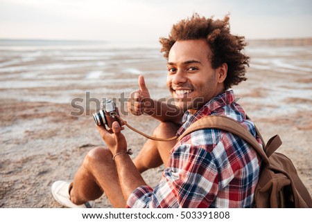 Happy african american young man with backpack taking photos and showing thumbs up on the beach