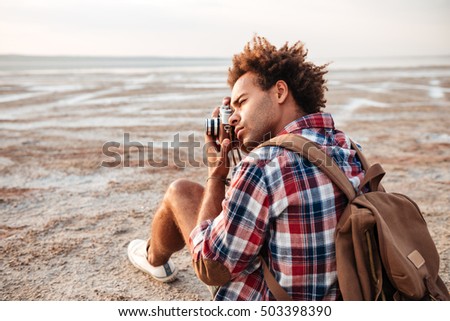 Concentrated african american young man with backpack sitting and taking pistures outdoors
