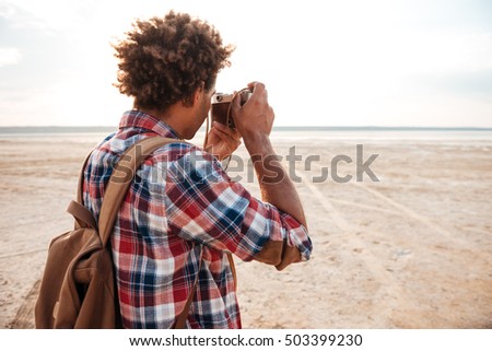 Back view of african american young man with backpack taking photos on the beach