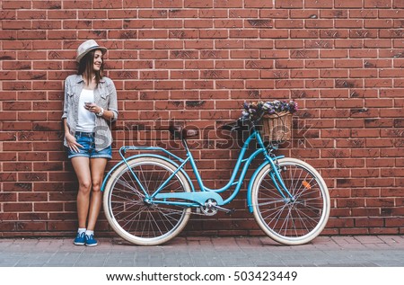 Beauty with vintage bike. Beautiful young smiling woman standing near her vintage bicycle with basket full of flowers while she leans against the wall. Royalty-Free Stock Photo #503423449