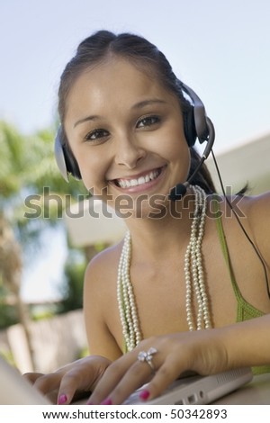 Young woman using laptop and headset at table in back yard, portrait