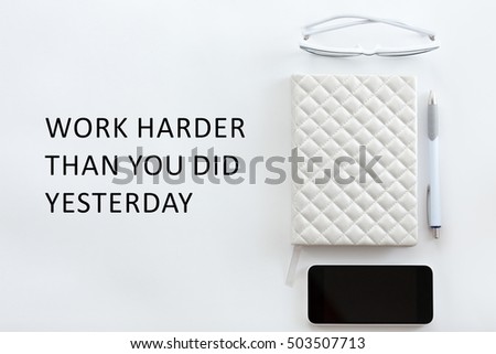 White office desk with glasses, mobile phone and pen on it. Top view. Motivational text "Work harder than you did yesterday"