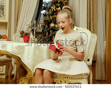 Excited blonde teenage girl in evening dress opening a gift box on Christmas.