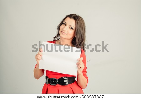 Beautiful young brunette woman in a red dress with a black belt holding a white sheet of paper on a gray backgroun. Mock Up