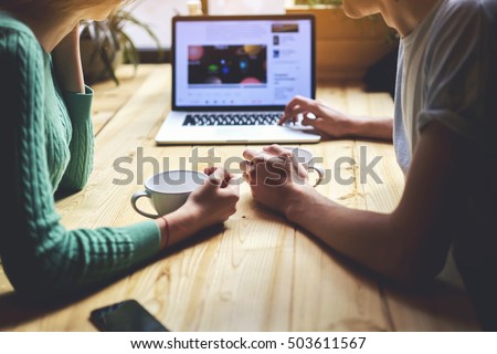 Group of friends are sitting at wooden coffee shop table with open laptop computer and two cups of tea. Couple of students are learning together by portable net-book. People watching video in internet Royalty-Free Stock Photo #503611567