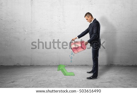 A businessman full-height in profile holding a red bucket with water pouring out of it on a green arrow pointing up, all on the background of a grey wall. Business and investment. Successful people
