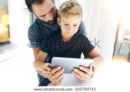 Father and his little son playing together on mobile computer, resting indoor.Bearded man with young boy using tablet PC in sunny home.Horizontal, blurred background, flares effect