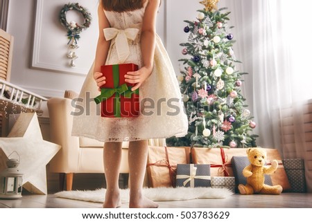Merry Christmas and Happy Holidays! Cheerful cute little child girl with present. Kid holds a gift box near Christmas tree indoors.