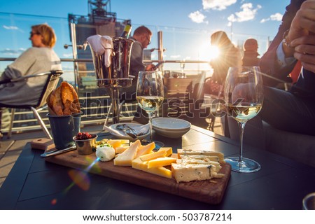 People eating cheese and drinking wine at rooftop restaurant at sunset time. Restaurant table served with cheese plate, bread and white vine full of visitors. Royalty-Free Stock Photo #503787214