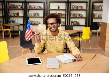 Cheerful african american young man holding united states flag sitting in library