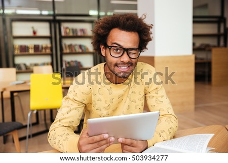 Happy cute african young man in glasses with tablet in library