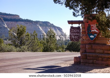 The wooden sign of Zion National Park with stone pillar and a big tree at east entrance on  Zion-Mount Carmel highway with Checkerboard Mesa backdrop in bright sky on sunshine day.
