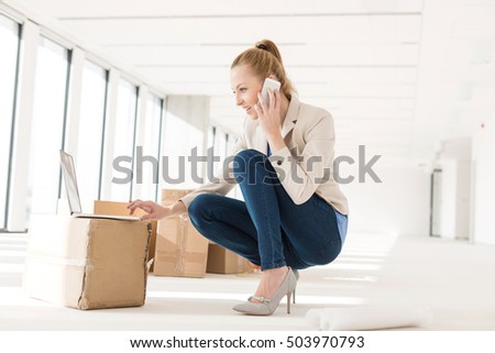 Full length of young businesswoman crouching while using mobile phone and laptop in new office
