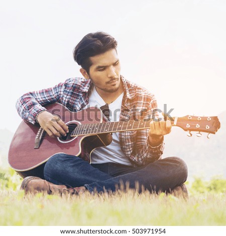 Young man playing acoustic guitar folk song, sitting on green grass. Nature background.