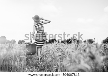 Black and white picture of woman in hat with suitcase walking away through wheat field. Back view of girl in striped dress in direct sunlight on summer countryside background.
