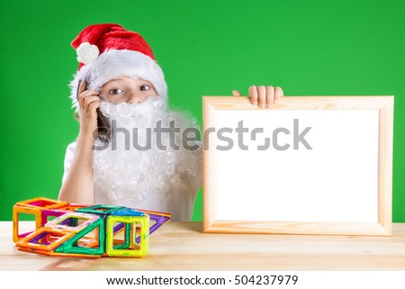 Little Santa talking on the phone, holding a blank picture frame with white background. Sitting at a wooden table. On the table lies a toy. He looks into the camera. Close-up. Green background.