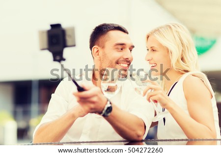love, date, technology, people and holidays concept - happy happy couple taking picture with smartphone on selfie stick and clinking glasses at city street cafe or restaurant