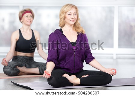 Fitness, stretching practice, group of two attractive fit happy senior females in sportswear working out in sports club, sitting in Easy (Pleasant Posture), Sukhasana, on meditation session in class Royalty-Free Stock Photo #504280507