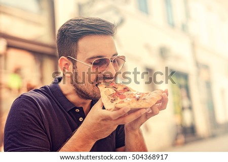 Handsome young man eating a slice of pizza outside on the street.