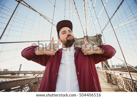 Handsome hipster guy taking selfie portrait on Brooklyn Bridge, New York. Cute bearded student takes funny picture with thumbs up for travel blog.