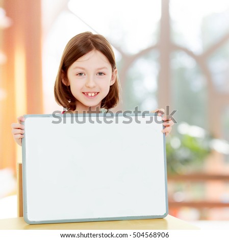 Cute little girl sitting at table holding in front of a white poster.In a room with a large semi-circular window.