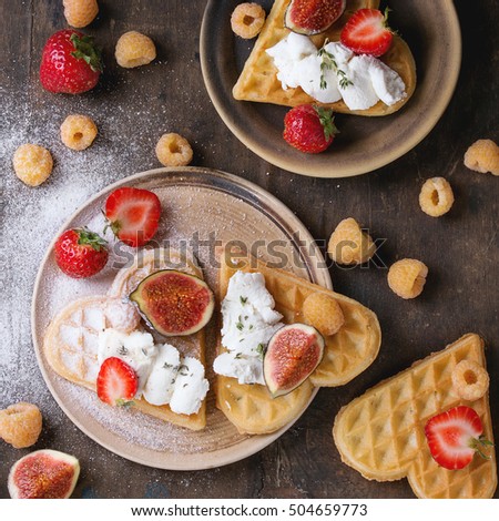 Wafers as heart shape with yellow raspberries, strawberries, sliced figs, ricotta cheese and sugar powder on ceramic plates over dark wooden background. Top view.  Square image