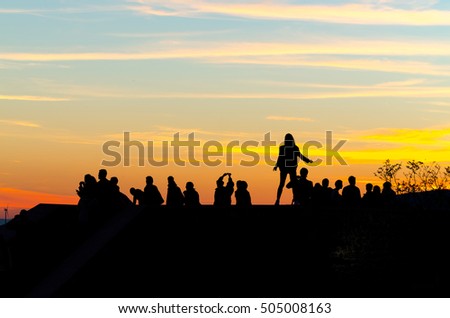 Silhouettes of young woman with long hair and people standing on a top of a view side taking photos of a colorful sunset sky with glowing clouds and  wind power plant in the background.