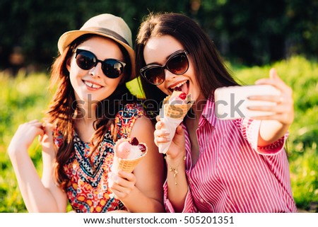 Portrait of two young women standing together eating ice cream and taking selfie photo on camera in summer street.