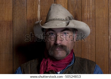 Close-up portrait of a cowboy with a mustache in front of a rough wood wall. He is staring at the camera with a serious expression. Horizontal format.