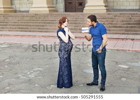Funny image. Couple expecting a baby: man holds a sign saying "boy?" Pregnancy in long dress. Future daddy in jeans and blue shirt.