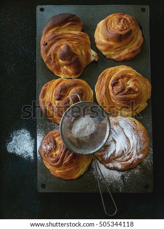 Fresh baked modern pastries cruffins, like croissant and muffin with sugar powder, served in silicon baking form with sieve over black background. Top view, natural day light