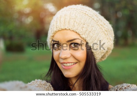 Closeup selfie of beautiful young Caucasian brunette woman in knitted beanie hat, smiling, outdoors in park. Pretty teenage girl taking a selfie in park in autumn. Mild retouch, natural light.