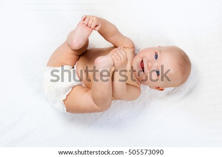 Funny little baby wearing a diaper playing on a white knitted blanket in a sunny nursery. Child after bath or shower on a fresh towel. Infant nappy change and skin care. Cute kid playing with his feet Royalty-Free Stock Photo #505573090