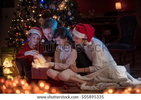 Christmas night. Near the christmas tree a lovely family opening their gifts. they are very happy to find a digital tablet inside. Mom and kids wearing a hat of Santa Claus.