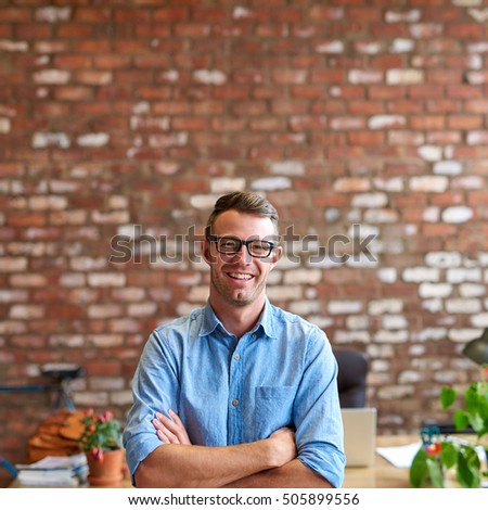Portrait of a casually dressed young businessman wearing glasses and standing in a trendy brick walled office