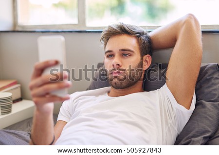 Handsome young man in pajamas using a cellphone while lying in bed in the morning
