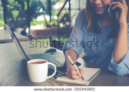 Beautiful businesswoman in casual style taking note and using smartphone and laptop on wooden table in coffee shop cafe with a cup of coffee.