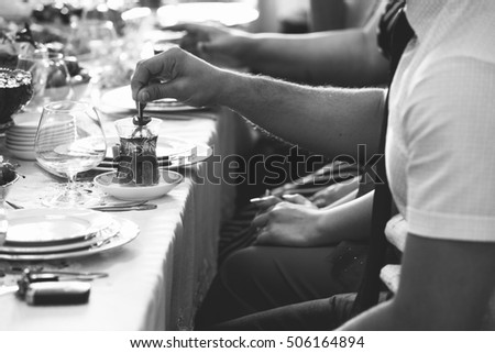Table setting of traditional tea in eastern glass with red ribbons on the white tablecloth, people mixing sugar in the tea with spoon, Arabic, Turkish, Azerbaijani customs, white and black