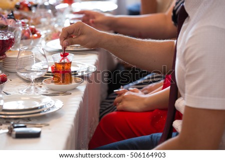 Table setting of traditional tea in eastern glass with red ribbons on the white tablecloth, people mixing sugar in the tea with spoon, Arabic, Turkish, Azerbaijani customs