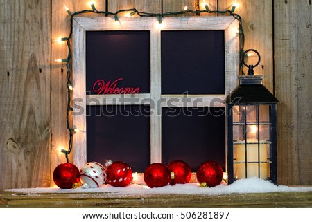 Blank rustic window frame with string of holiday lights, Welcome, antique lantern with candle, snow, and big red and silver Christmas ornaments by antique rustic wood background; black copy space 