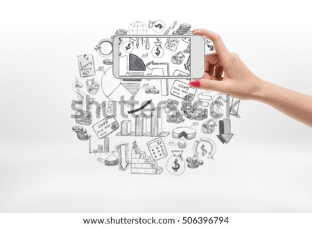 A female hand with a cellphone taking a photograph of a collage with a lot of financial icons and diagrams, on the white background. Business and finance. Success and profit. Infographics and analysis