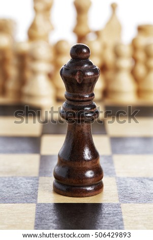 The photograph shows a still life of one of the pieces of the game made of dark wood in a vertical position standing on the checkered board with the white pieces in the backgound.