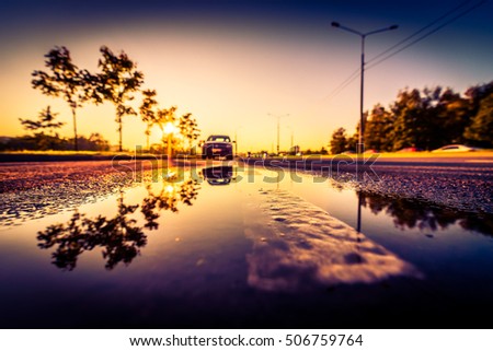 Sunset after rain, the car parked on the roadside and the headlights of the approaching cars. Wide angle view of the dividing line level in a puddle, image vignetting and the orange-purple toning